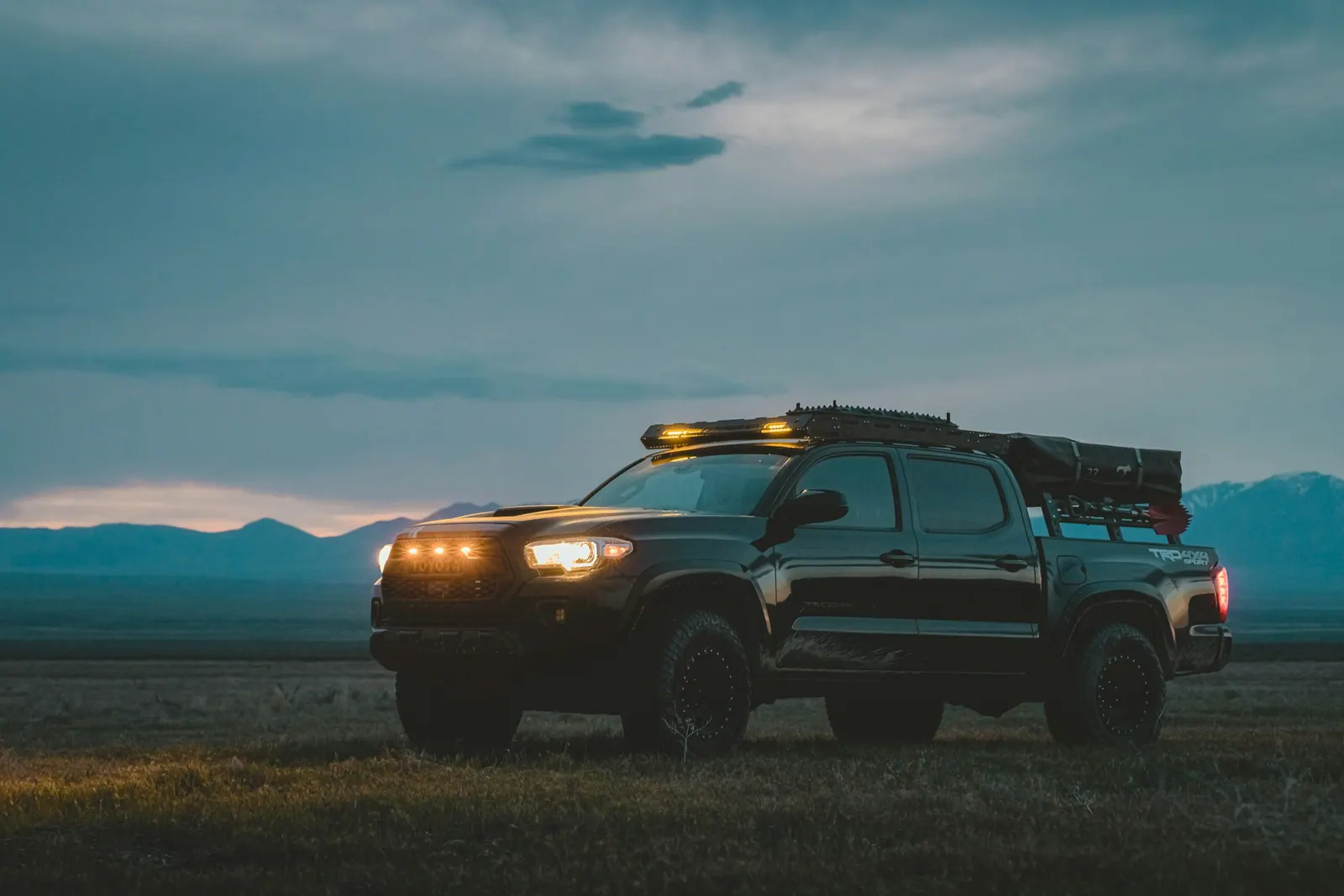 Dark pickup truck with a roof rack and its lights on, parked on open ground below mountains at dusk